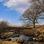 A stream flows over rocky terrain through a winter landscape in Wales, with bare trees silhouetted against blue skies and white clouds. Stone ruins from the historic Ynys y Pandy slate mill are visible, surrounded by moorland and distant hills.